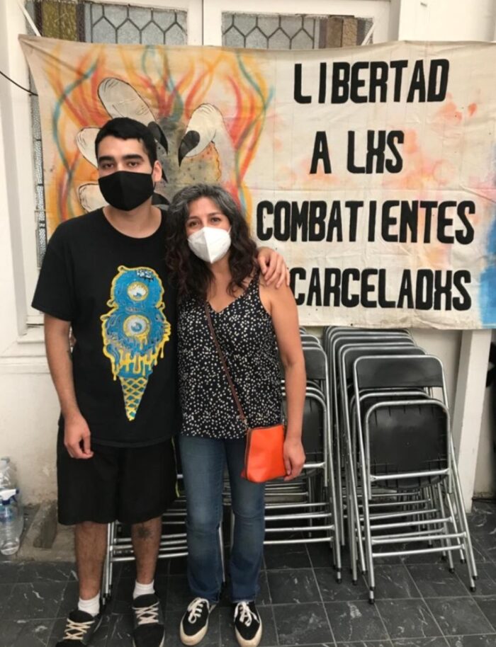 Óscar Pérez Cortez, sobreviviente del atropello por parte de Carabineros en 2019, junto a su madre en una actividad. Detrás de ellos hay un lienzo que dice ''libertad a les combatientes encarcelades''.