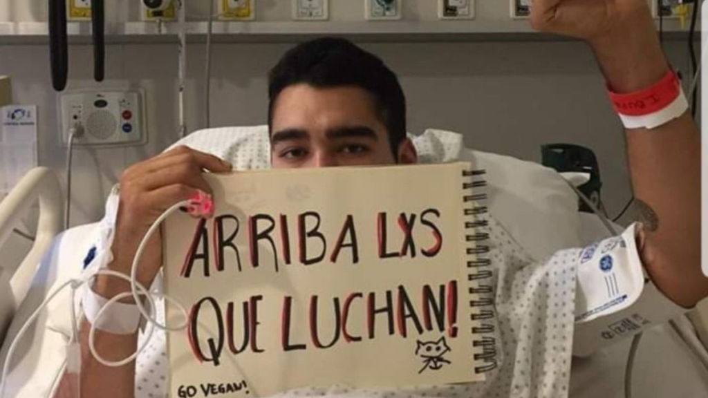 Óscar Pérez, víctima del atropello efectuado por Carabineros en Plaza Dignidad durante 2019. El joven está en una camilla de hospital y en su mano sostiene un cuaderno que dice ''arriba les que luchan''. En letras pequeñas y abajo de la frase, dice ''go vegan'' junto al dibujo de un gato. 