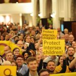 Grupo de personas manifestándose en el Día de la Intersexualidad en 2018. Llevan banderas de la intersexualidad y carteles que dicen "protejan a la niñez intersexual".