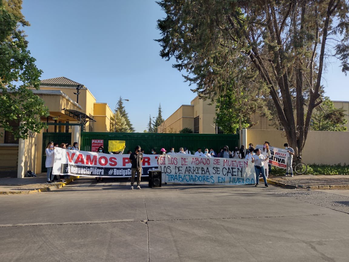 Imagen de trabajadoras y trabajadores de la chocolatería en una movilización por mejores condiciones laborales. Tienen tres lienzos, el más visible dice ''si los de abajo se mueven, los de arriba se caen. Trabajadores en huelga''. En el lienzo de atrás, se ven los hashtags #RenunciaPiñera y #HuelgaGeneral. El cielo es azul y detrás de los manifestantes hay una gran reja verde oscuro que resguardan a edificios color amarillo claro.