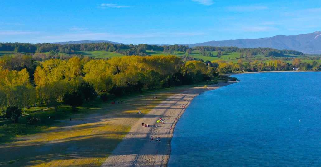 La fotografía esta toamada desde la altura. Del lado derecho aparece el lago ranco con un azul fuerte. Al medio esta la playa de arenas blancas, se ven a la distancia muy pequeños unos turistas. Del lado izquierdo esta el bosque, iluminado por la luz de la tarde. En ese lugar se pretende instalar el proyecto inmobiliario