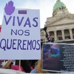 A la izquierda: cartel blanco con una mano morada y letras moradas que dicen "Vivas nos queremos". A la derecha arriba un edificio blanco antiguo y abajo un cartel con una mujer y letras blancas.